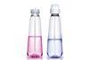 Two unlabelled, transparent plastic aerosol bottles sit side by side, containing pink and blue liquid, respectively. The one on the left is missing its nozzle, displaying the valve. The one on the right has the nozzle attached.