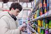A man wearing headphones, stood in a supermarket isle, looking at a can of energy drink