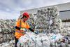 A worker wearing a safety helmet and hi-vis jacket stands amongst towering piles of plastic bales. She is reaching out to touch one.