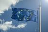 A European Union flag is waving in the wind on a flagpole. The sky behind it is blue, with a few clouds on the left hand side of the image and sunlight beaming down from the top right corner.