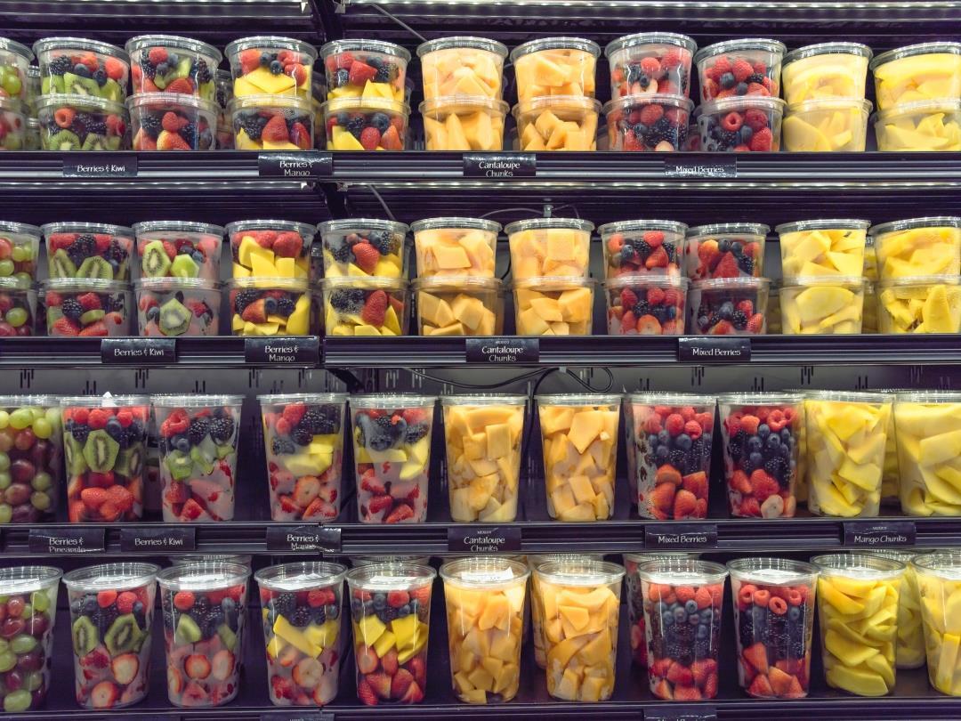 Four supermarket shelves are lined with plastic tubs full of chopped fruits. From left to right, each shelf displays the…