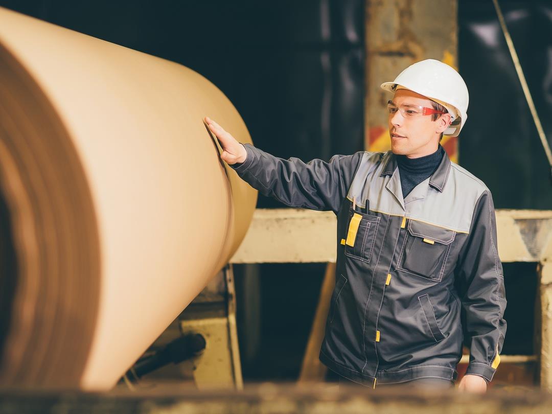 An employee in a paper factory places his hand on a giant roll of paper. He is wearing a protective jacket and hard hat.