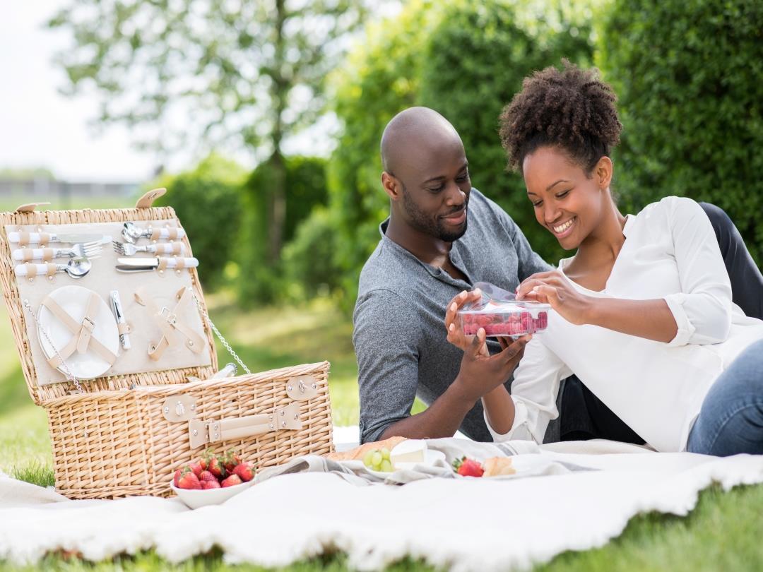 A heterosexual couple lay on a picnic blanket in a park. The woman is peeling open a plastic tray full of raspberries. An…
