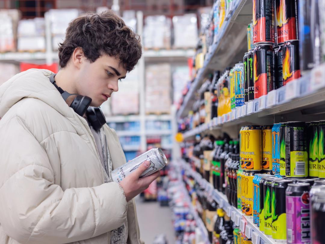A man wearing headphones, stood in a supermarket isle, looking at a can of energy drink