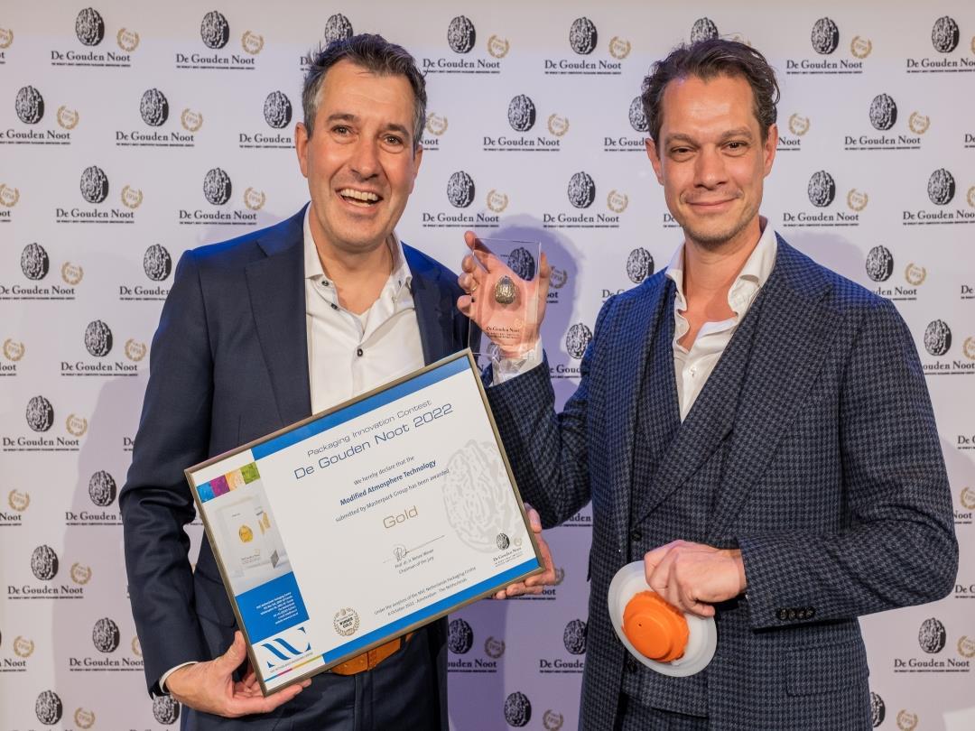 Two men wearing suits smile at the camera. The man on the left is holding a transparent trophy with a solid gold walnut…