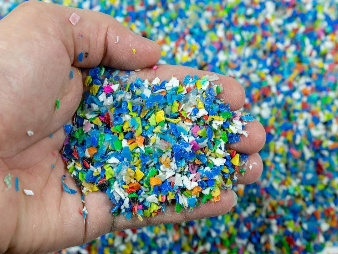 A pile of colourful polymer pellets viewed from above. A hand has scooped up some of the flakes and is holding them up…