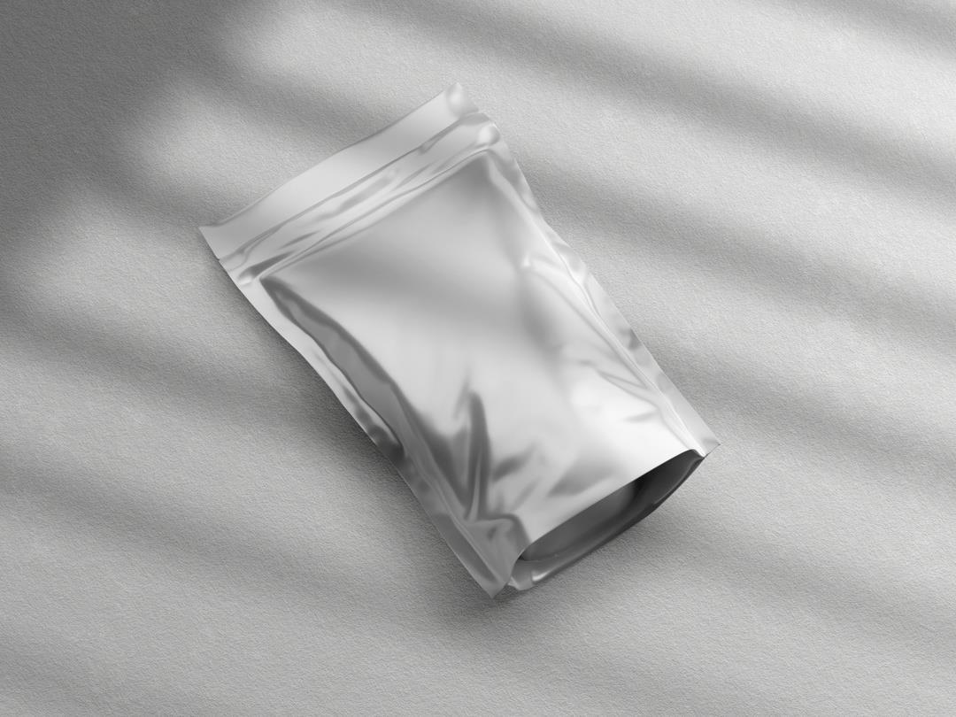 A 3D render of a flexible pouch. It appears to be made of a shiny silver foil, and is laying on a rough white surface.…