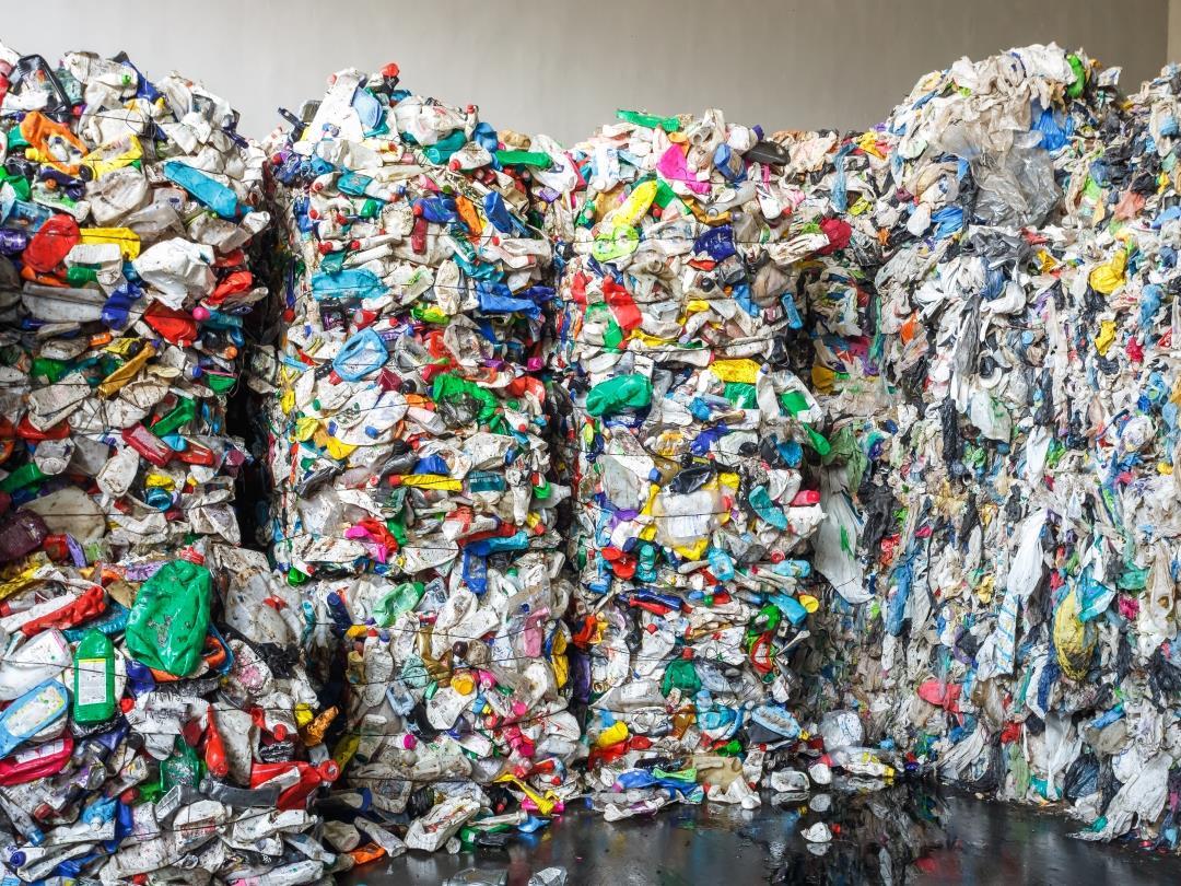 Four large bales of recycled plastic packaging. Various bags, bottles, and tubs have been crushed and tied together at a…