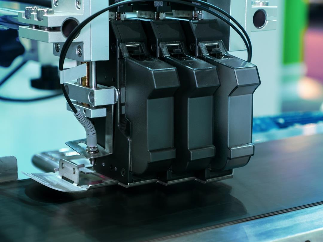 A close-up photo of an industrial inkjet printer on a conveyor belt. Attached to a mechanical arm, it has three ink…