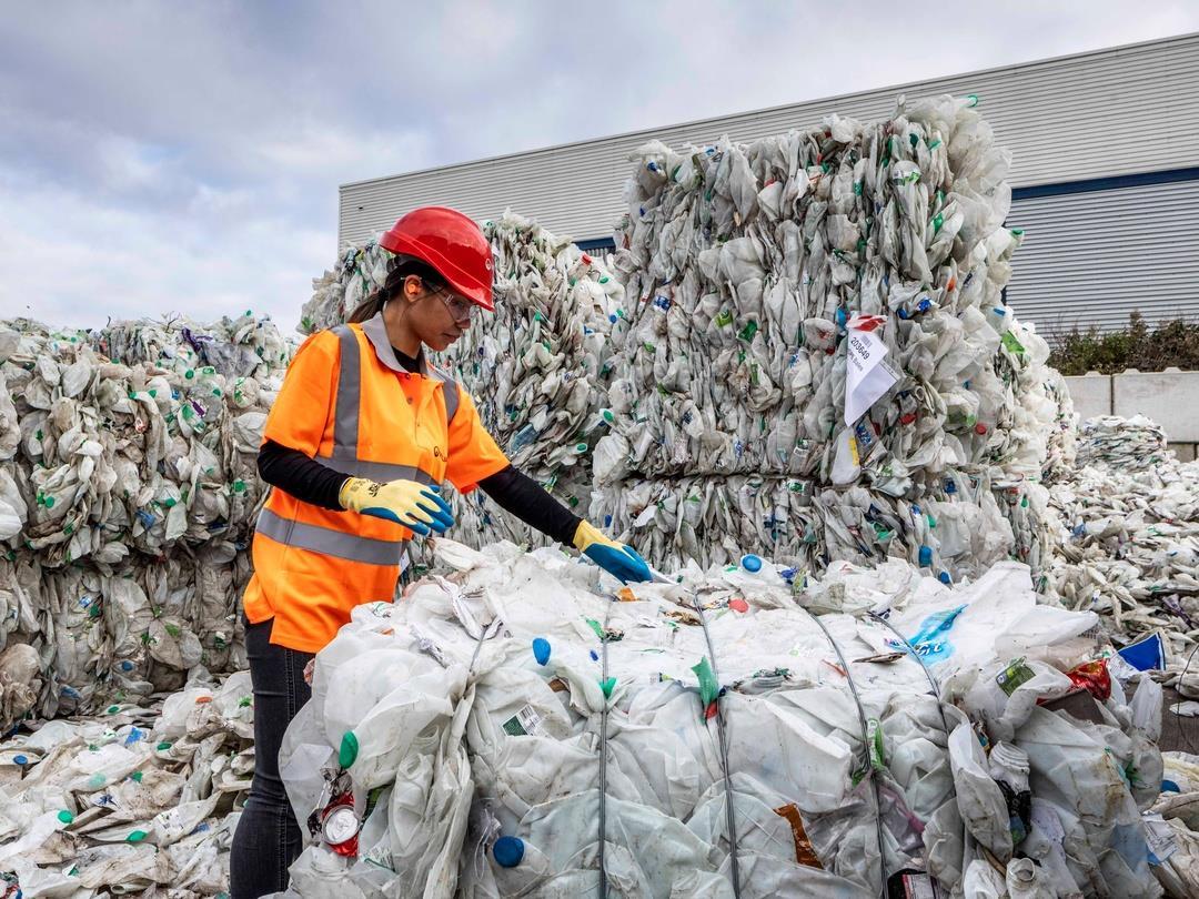 A worker wearing a safety helmet and hi-vis jacket stands amongst towering piles of plastic bales. She is reaching out to…