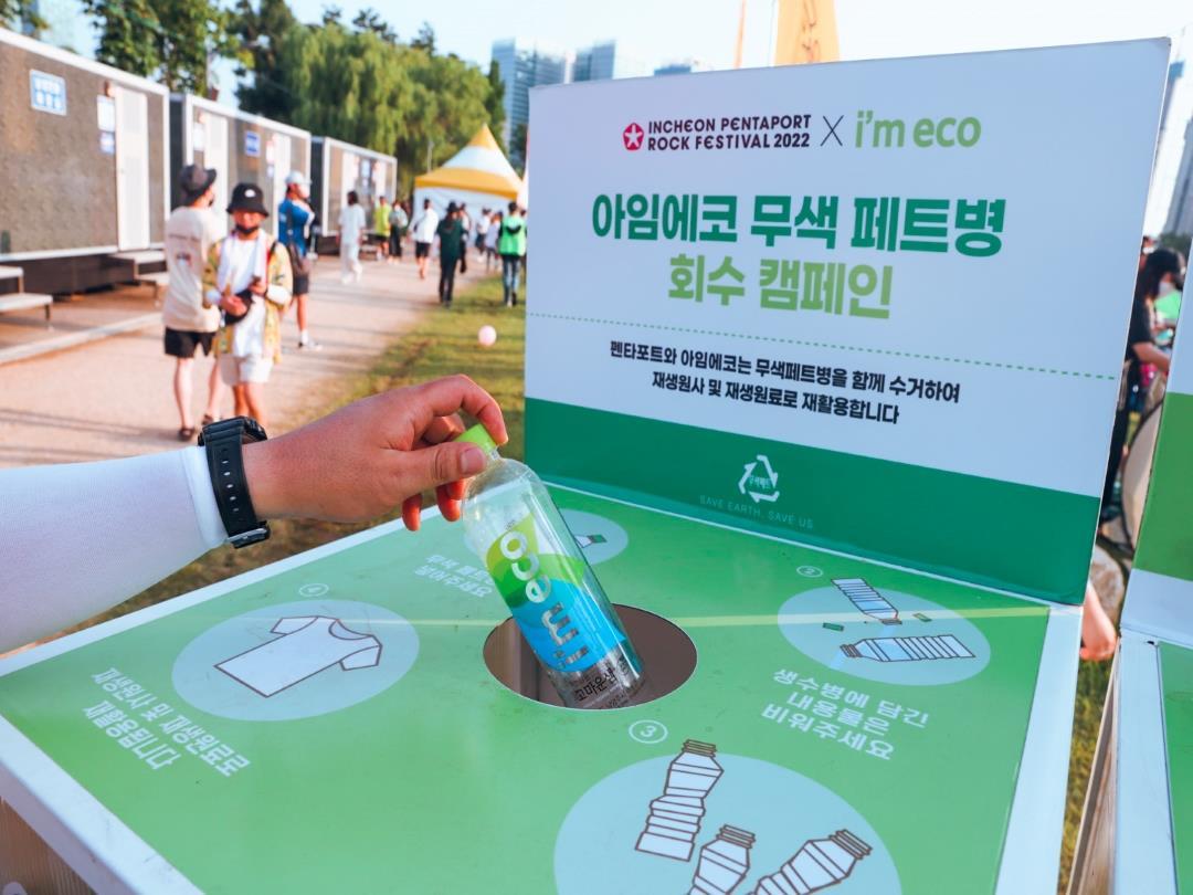 A hand extends from off-camera, holding a bottle labelled 'I'm Eco'. It is being held over a recycling bin, on which is…