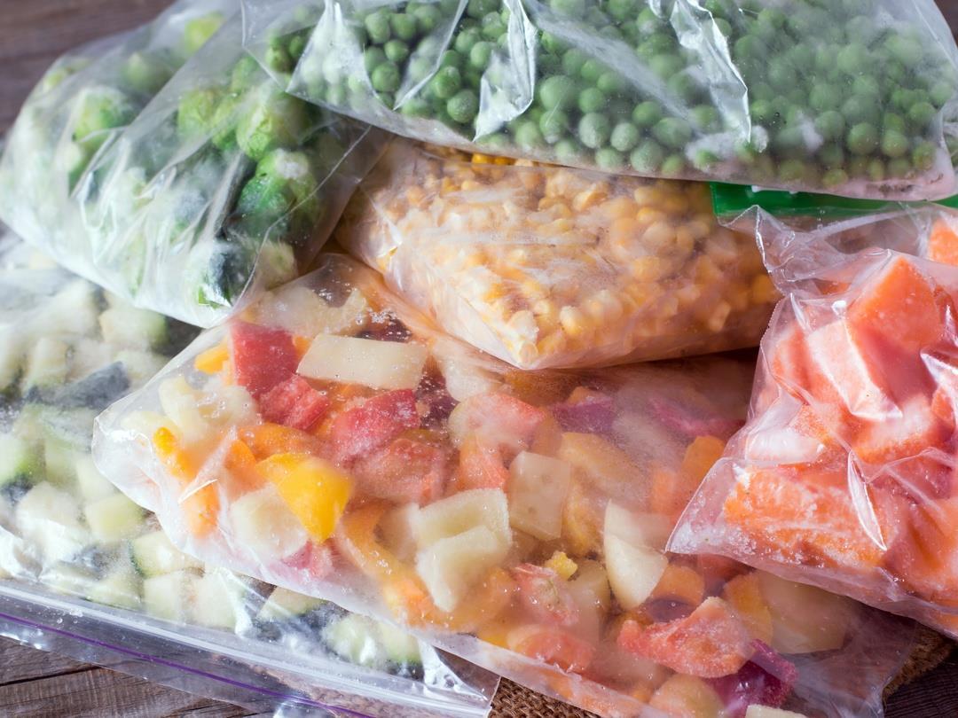 Bags of frozen vegetables, including peas, Brussels sprouts, and chopped carrots, lay in ziploc plastic bags on top of a…