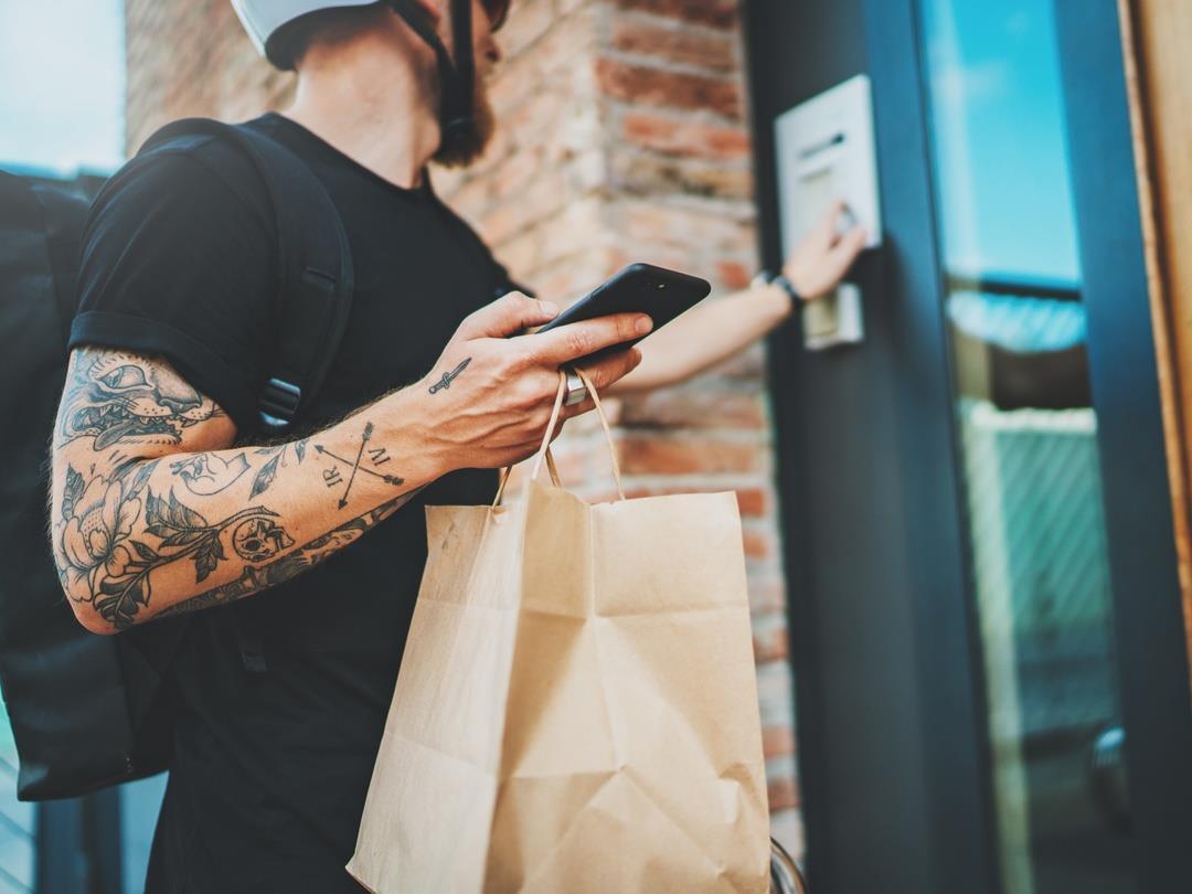 A delivery man holds a paper bag up to the camera. He appears to be ringing a doorbell at an apartment block, although this…
