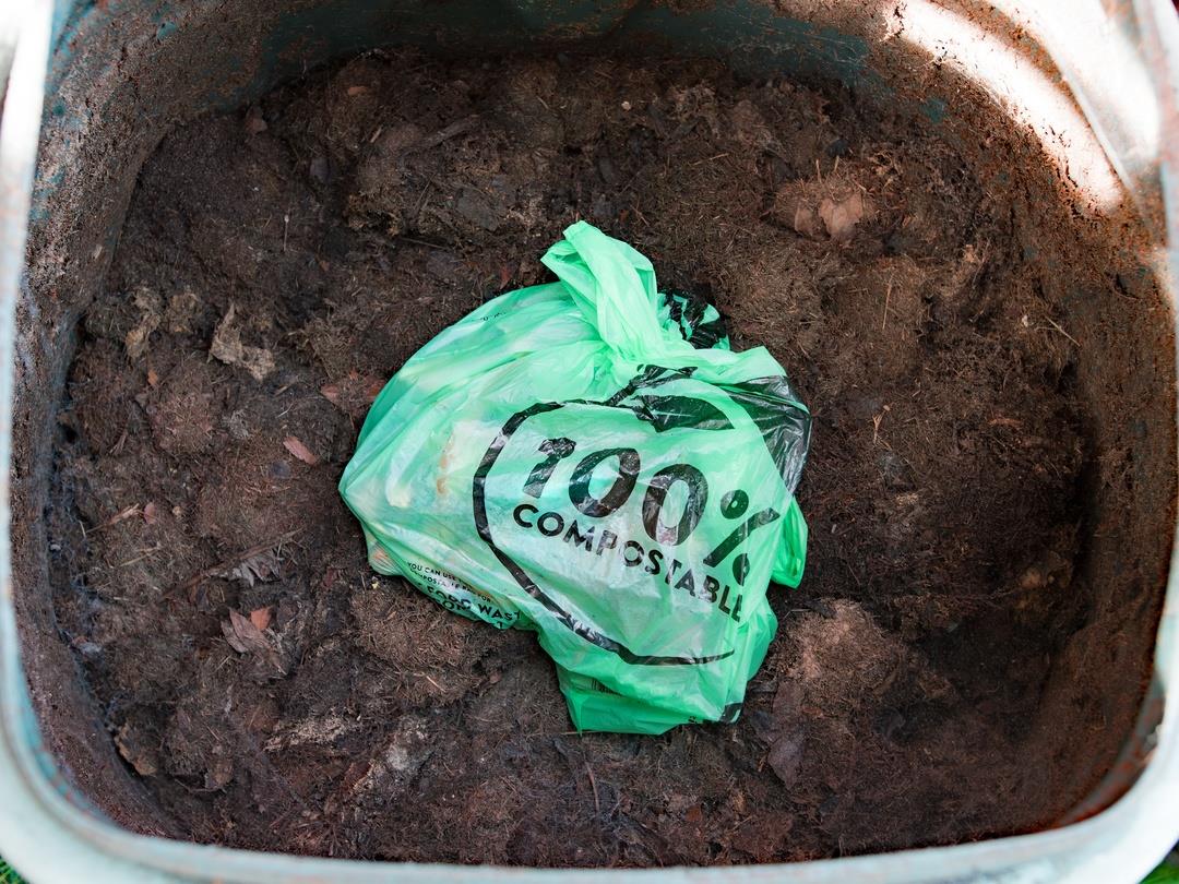 The camera looks down into an open compost bin. On top of the compost sits a light green plastic bag, tied at the top,…