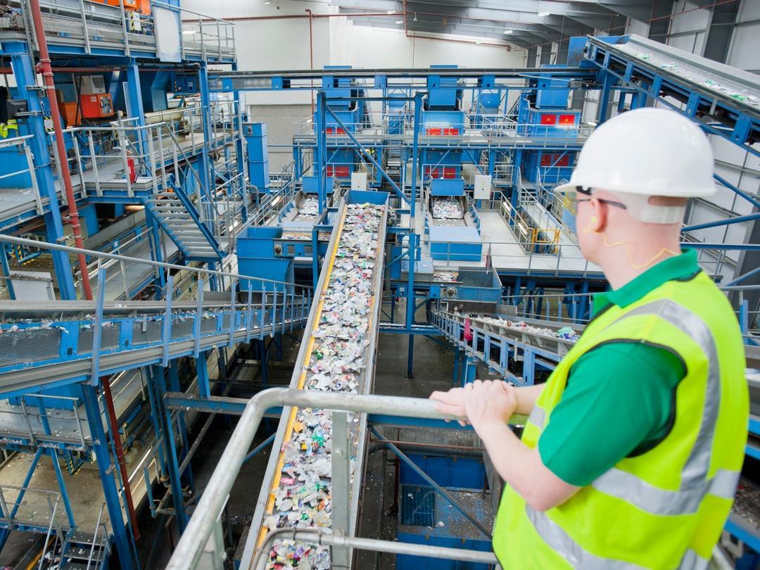 A worker at a recycling plant, wearing a hard hat and hi-vis jacket, is standing over a conveyor belt carrying plastic…