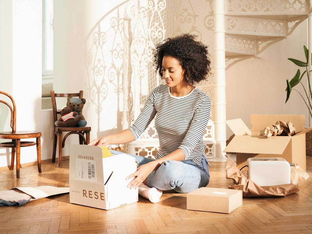 A woman sits cross-legged on her living room floor. She is opening a cardboard box. Other e-commerce packaging is scattered…