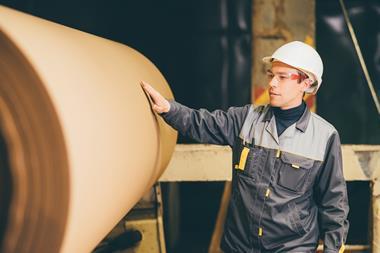 An employee in a paper factory places his hand on a giant roll of paper. He is wearing a protective jacket and hard hat.