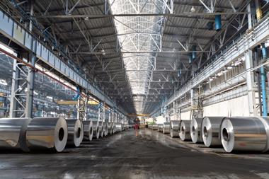 An interior shot of the production shop at an aluminium plant. Rolls of aluminium are placed in rows extending away from the camera, creating an aisle; a forklift drives through it in the distance.