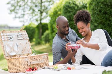A heterosexual couple lay on a picnic blanket in a park. The woman is peeling open a plastic tray full of raspberries. An open picnic basket and bowls of strawberries and grapes are placed nearby.