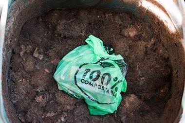 The camera looks down into an open compost bin. On top of the compost sits a light green plastic bag, tied at the top, featuring black text that reads, '100% compostable'.