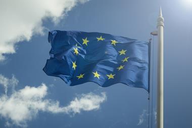 A European Union flag is waving in the wind on a flagpole. The sky behind it is blue, with a few clouds on the left hand side of the image and sunlight beaming down from the top right corner.