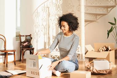A woman sits cross-legged on her living room floor. She is opening a cardboard box. Other e-commerce packaging is scattered around her, including a paper pouch containing clothes, unwrapped paper revealing a toaster, and an unopened cardboard box.