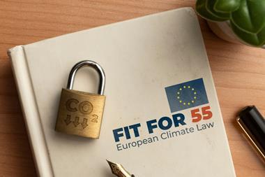 A white hardback notebook lays on a wooden table. On the cover is the EU flag and the words 'FIT FOR 55 European Climate Law'. On top of it lays a padlock with the phrase 'CO2' and downward-pointing arrows engraved into it.