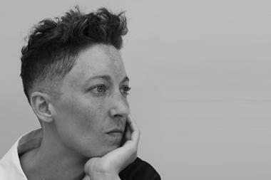 Black and White headshot of a white queer person with olive, freckled skin. Short dark fade, curly on top. Light eyes peer into the distance, as she rests her chin on her hand