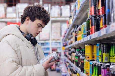 A man wearing headphones, stood in a supermarket isle, looking at a can of energy drink