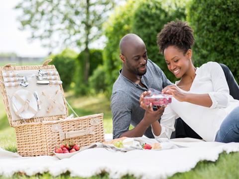 A heterosexual couple lay on a picnic blanket in a park. The woman is peeling open a plastic tray full of raspberries. An open picnic basket and bowls of strawberries and grapes are placed nearby.