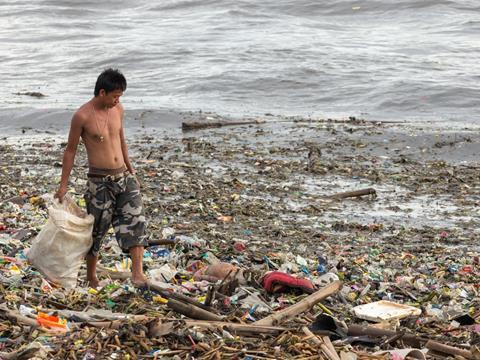 A man is walking across a severely polluted seafront. He is holding a bag and collecting the waste.