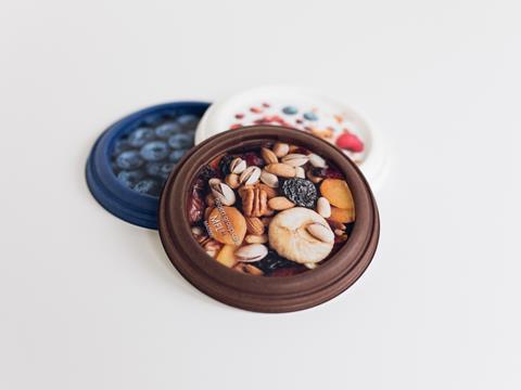 An assortment of circular moulded fibre lids lays on a white surface. The lid closest to the camera is brown, with a picture of various nuts printed onto it. Small text on the lid reads ‘hebert-group.com, MFL’.