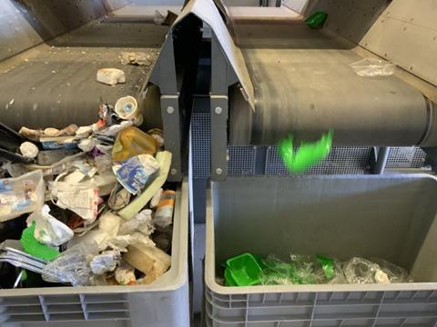 A photograph of two separated waste streams, with two conveyor belts dropping waste into boxes. The stream on the right is sorting PLA bottles and trays into one box. Other waste, including plastic bags, trays, and paper cups, are on the left.