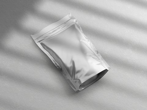 A 3D render of a flexible pouch. It appears to be made of a shiny silver foil, and is laying on a rough white surface. Sunlight appears to be shining through a pair of shutters, casting rectangles of light over the image.