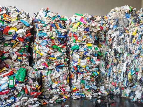 Four large bales of recycled plastic packaging. Various bags, bottles, and tubs have been crushed and tied together at a processing plant.