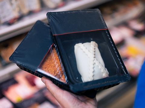 A hand is holding two plastic food trays containing breaded meat and an uncooked pastry. Each tray is lidded with a transparent film. There are supermarket shelves in the background, although they are out of focus.