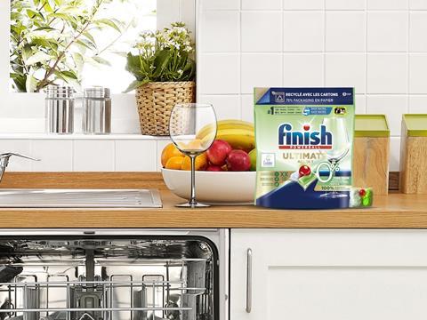 A photo of a kitchen counter above an open dishwasher. On its surface sits a clean wine glass next to a pouch of Finish dishwasher capsules. The packaging claims that it is ‘Recyclé avec les cartons’ and ‘75% packaging en papier’.