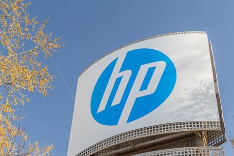 The camera looks up at a large HP sign from below. Behind it, the sky is blue, and a tree with orange blossoms is visible.