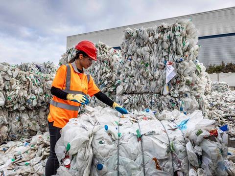 A worker wearing a safety helmet and hi-vis jacket stands amongst towering piles of plastic bales. She is reaching out to touch one.