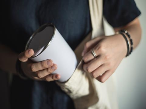 A close-up on a person's shoulder. They are wearing a tote bag, out of which they are pulling a reusable metal coffee cup.