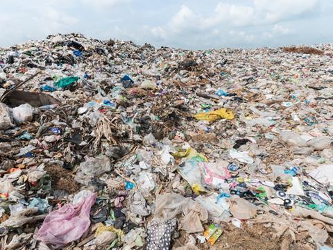 A large pile of landfill towers above the camera, containing waste such as plastic bags, crushed bottles, food packaging, and bin liners.