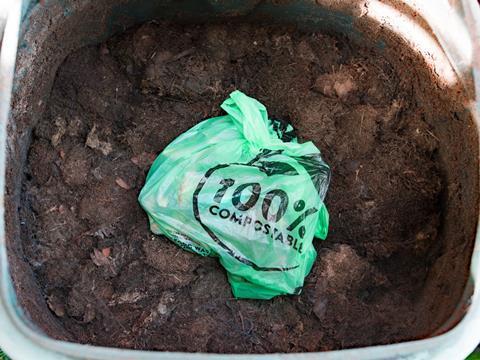 The camera looks down into an open compost bin. On top of the compost sits a light green plastic bag, tied at the top, featuring black text that reads, '100% compostable'.