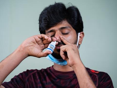 A man has tucked his medical face mask under his chin to access his nose. Pressing down on his left nostril, he has inserted a nasal spray into his right. The packaging reads, 'Intranasal COVID-19 vaccine'.
