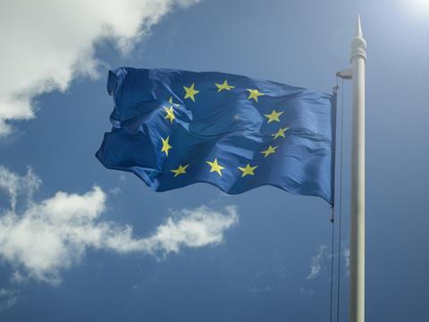 A European Union flag is waving in the wind on a flagpole. The sky behind it is blue, with a few clouds on the left hand side of the image and sunlight beaming down from the top right corner.