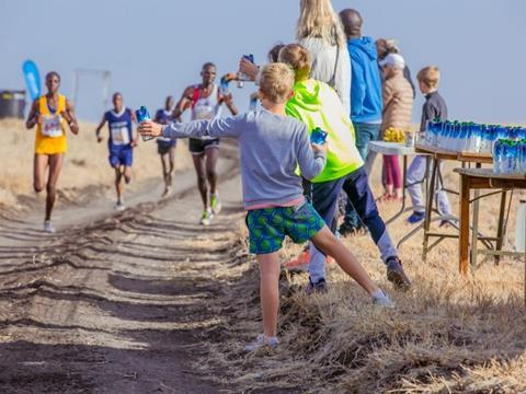 Runners on a marathon track in Kenya. To the left of the track, a table has been set up with paper Tetra Pak cartons full of water. Spectators are holding them out for the runners to grab as they pass.