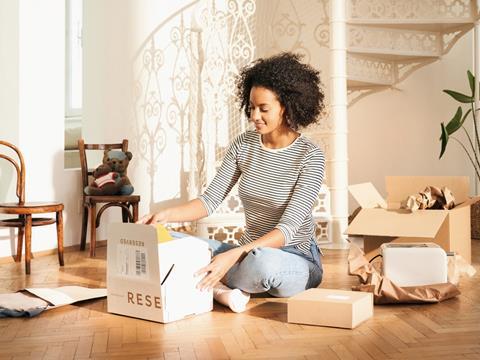 A woman sits cross-legged on her living room floor. She is opening a cardboard box. Other e-commerce packaging is scattered around her, including a paper pouch containing clothes, unwrapped paper revealing a toaster, and an unopened cardboard box.