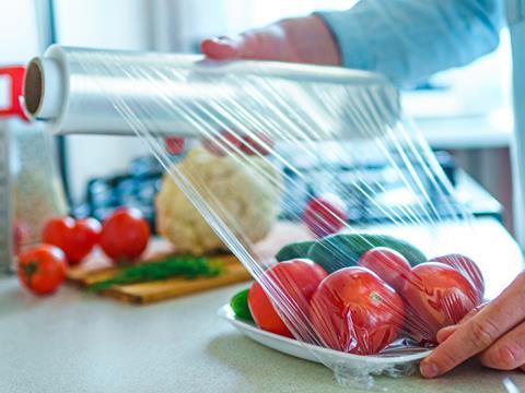 A man is wrapping a plate of tomatoes - and another green vegetable, although it is blurred out by the camera focus - in cling film.