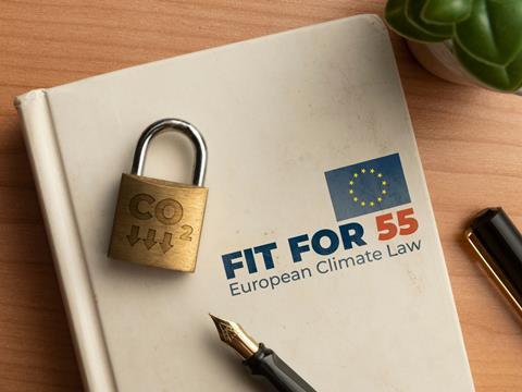 A white hardback notebook lays on a wooden table. On the cover is the EU flag and the words 'FIT FOR 55 European Climate Law'. On top of it lays a padlock with the phrase 'CO2' and downward-pointing arrows engraved into it.