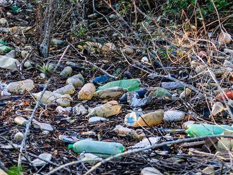 Plastic bottles have been littered amongst the bark, twigs, and weeds on a woodland ground.