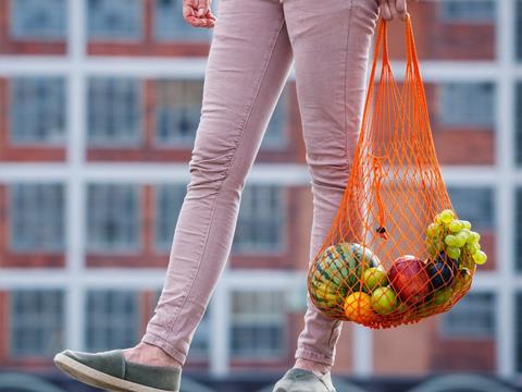 A woman's legs against the backdrop of a block of apartments. She is holding a mesh bag containing fruits and vegetables such as a watermelon, a bunch of grapes, and an orange.